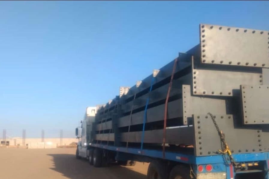 Plataforma cargada con estructuras de acero de gran formato, aseguradas con bandas y cadenas.
