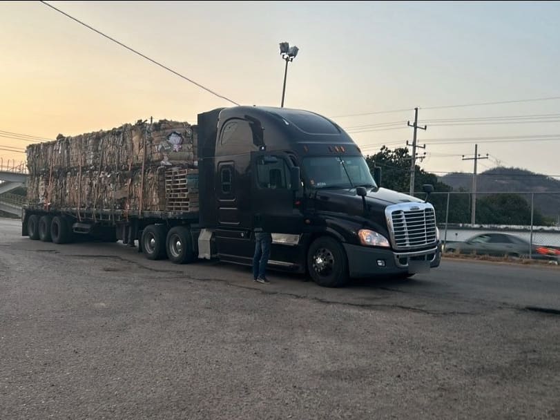Tractocamión con plataforma cargada de material enfardado, listo para salir a carretera al atardecer.